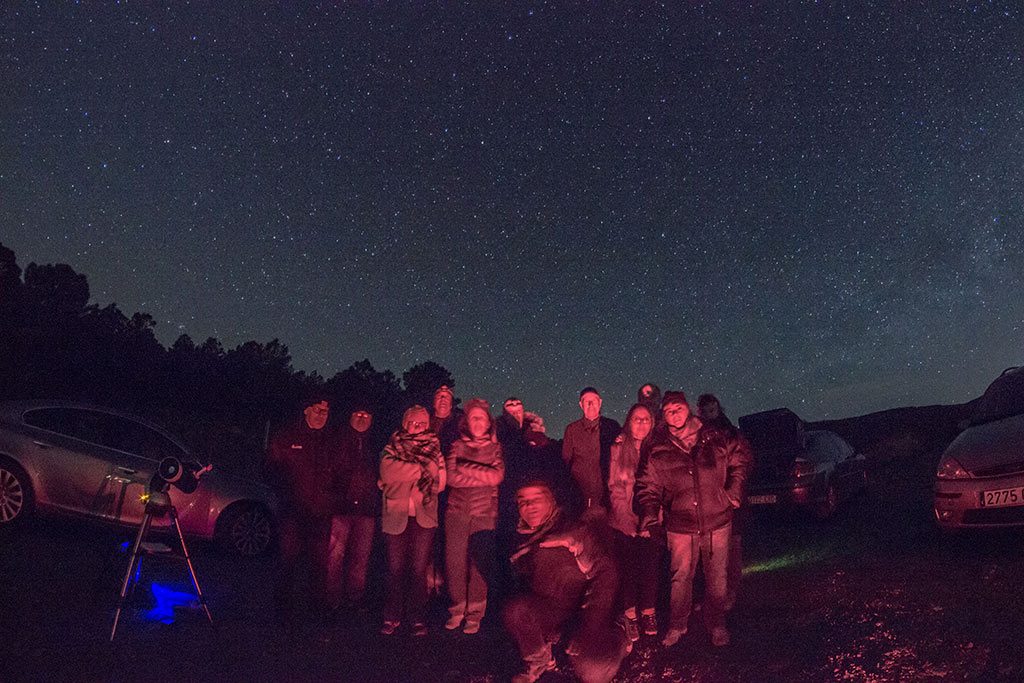 Grupo de personas durante una observación astronómica. Aparece un grupo de hombres y mujeres muy abrigados, bajo un cielo estrellado. Hay también algún telescopio. Están iluminados solo por la tenue luz de una linterna. Disfrutan del cielo estrellado.